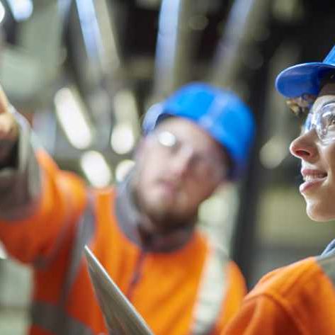 two industrial service engineers conduct a safety check of a control panel and boiler room at a power station . They are both wearing safety equipment and are looking at a control panel .